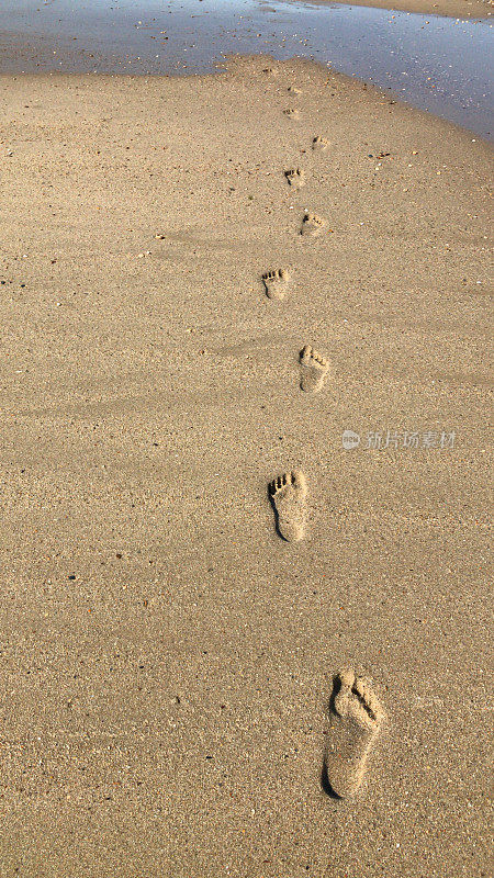 海滩脚印在沙滩上的图像，赤脚/光脚脚趾在海边沿着英国海岸线的海水边缘，低潮夏天日落的脚，脚印印象3d光学错觉，一个人行走在沙漠海滩度假